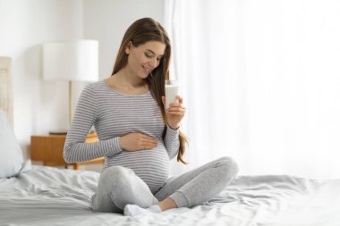 Avrupalı hamile bir kadın, rahat bir evde, bir yatakta rahatça oturuyor, bir bardak süt tutuyor, hamilelik boyunca mutluluk ve huzur saçıyor.