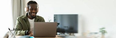 Kulaklıklı mutlu Afro-Amerikan adam dizüstü bilgisayarla çalışıyor, ev ofisinin masasında oturuyor ve internet üzerinden müşterilere danışmanlık yapıyor.