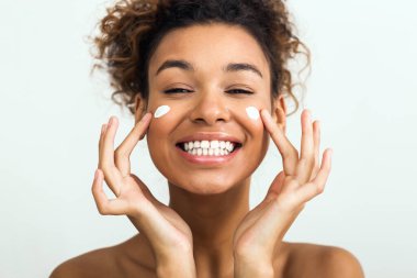 A cheerful African American lady with a glowing smile applies facial cream, symbolizing skincare and self-care routines clipart