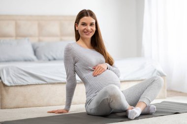 Gülümseyen hamile bir kadın yoga minderinde rahat bir şekilde oturur. İyi aydınlatılmış bir yatak odasında, sakinlik ve pozitif titreşimler yayar.