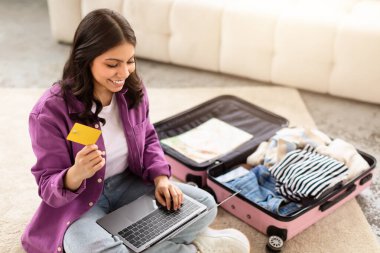 Genç bir ortadoğulu kadın, açık bavulunun yanında kredi kartı ve dizüstü bilgisayarla seyahat için hazırlanırken görülüyor.