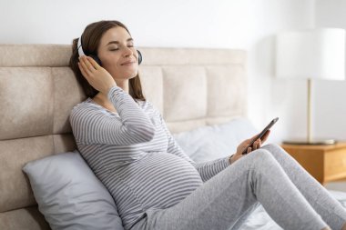 Huzurlu hamile bir kadın kulaklıklarla müzik dinlemekten zevk alıyor, yatakta rahat bir şekilde oturuyor, akıllı bir telefon tutuyor.