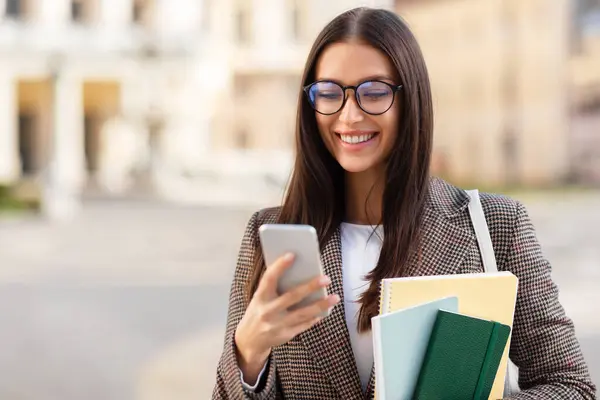 Intelligent young woman student with eyeglasses texts on phone, academic building in the background
