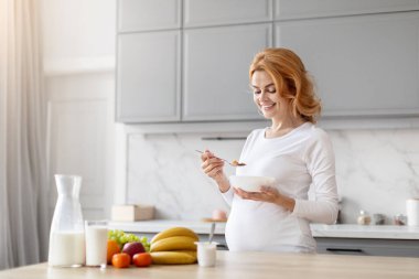 Avrupalı hamile bir kadın sağlıklı bir kahvaltı yapıyor, doğum öncesi beslenme ve güneş gören bir mutfak ortamında refahı vurguluyor.