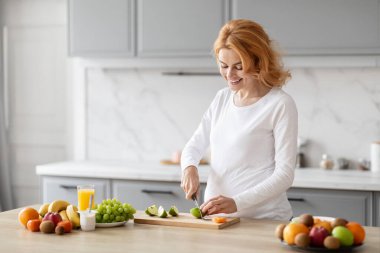 Parlak bir mutfakta Avrupalı hamile kadın sağlıklı yaşam tarzını betimlerken taze meyve kesmeye odaklanmış.