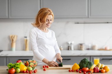 Gülümseyen hamile bir kadın, taze sebze ve meyvelerle çevrili modern bir mutfakta salatalık dilimliyor.