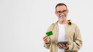 A senior man is seen isolated on white, looking at his tablet and holding a credit card, representing the elderly adapting to modern technology and online banking, copy space clipart