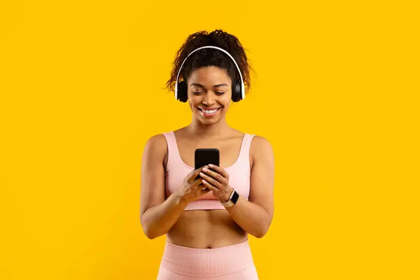明るい黄色の背景に彼女のスマートフォンを使用してスポーティな服で若いアフリカ系アメリカ人女性を笑う — ストック写真