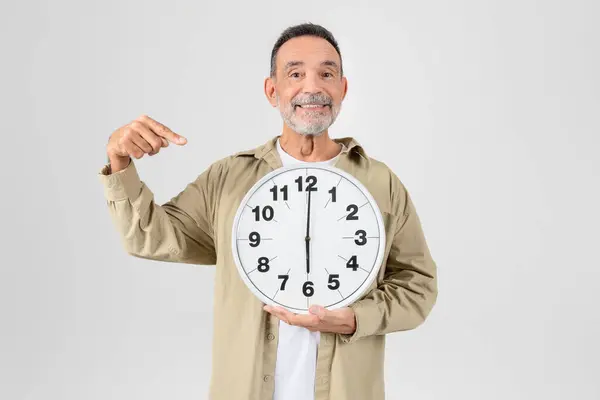 ひげのある陽気な老人が大きな壁時計を持ち それを指差し 明白な背景に立っています — ストック写真