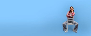 Gündelik giysiler içinde gülümseyen Asyalı bir kadın, metin veya tasarım elementleri için yeri olan mavi bir zemine karşı bir sandalyede oturur.