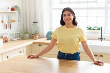 Neşeli, gülümseyen bir Arap kadın, sıcak ve davetkar bir atmosfer sunarak, kendi mutfağında güvenle duruyor.