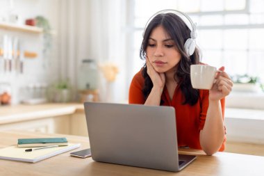 Kulaklık takan orta doğulu bir kadın, kahve içerken ekrana odaklanıyor ve online ders dinliyor.