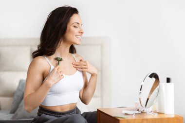 Beyaz bluzlu, gülümseyen bir kadın, yeşil yeşim bir silindir kullanıyor, yatak odasında ayna ve ürünlerle oturuyor.