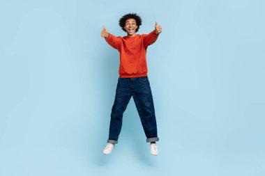Enerjik afro-amerikan adam havada sıçrıyor, iki parmağını havaya kaldırıyor, durdurulamaz neşe ve pozitif titreşimleri izole edilmiş mavi bir zeminde ifade ediyor.