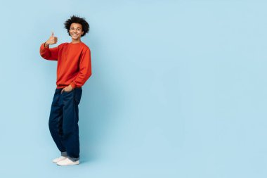 Neşeli, Afro-Amerikan bir adam, sıradan giyinmiş, pozitiflik saçan, pozitiflik saçan, pozitifliği taklit eden, mavi bir arka plana başparmağıyla poz veriyor.