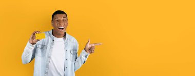 Bir kredi kartı gösteren neşeli bir adam ve heyecan verici bir şekilde web-model sarı bir arka planda geniş bir boşluğa işaret ediyor.