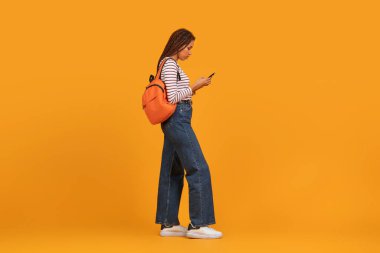 Serene 'li genç siyahi kadın, turuncu arka planda izole edilmiş uluslararası cazibe ve çok ırklı moda eğilimleri sergileyerek modern bir atmosferle yürüyor.