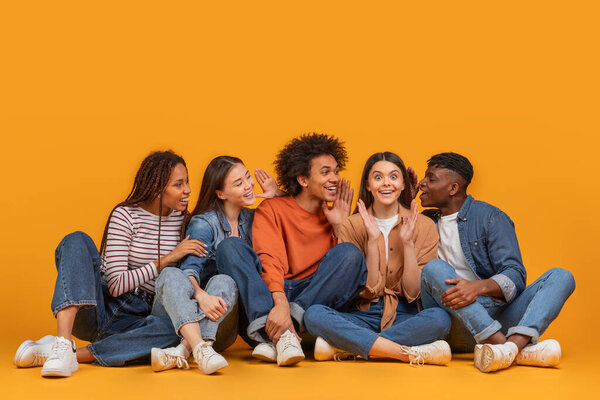 Young international friends share whispers and smiles, capturing a moment of friendship in a multiracial, multiethnic scenario, isolated on a yellow background