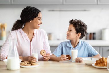 Bu görüntü, Afrika kökenli Amerikalı anne ve çocuğu kurabiye yerken yakalıyor, sıcak bir aile ortamını sıcak bir mutfak ortamında paylaşıyor.