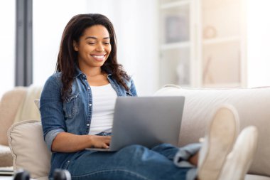 Mutlu Afro-Amerikan kadın bir kanepede oturur, dizüstü bilgisayarına dalmış bir şekilde. Yazıyor ve kayıyor, işine ya da bilgisayardaki faaliyetlerine odaklanıyor..