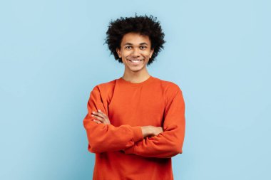 Afro-Amerikan genç bir adam kollarını kavuşturarak güven ve yakınlık yayıyor, turuncu bir kazak giyiyor, koyu mavi izole bir arka plana karşı duruyor.