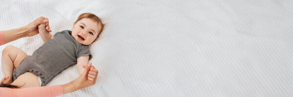 Mother care. Unrecognizable mommy holding hands of her smiling little baby, caring mom bonding with infant boy or girl while child lying on bed at home, free copy space, web-banner