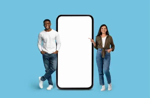 stock image Smiling Multiracial couple pointing towards a huge mobile phone illustration on blue studio background