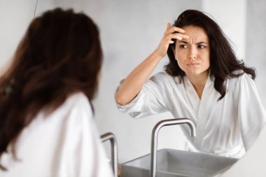 A woman in a white robe appears concerned as she examines her forehead for wrinkles in the bathroom mirror, embodying beauty anxieties clipart