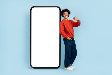 Genç, neşeli, sıradan kıyafetler giyen Afro-Amerikalı bir adam boş ekranlı dev bir akıllı telefonun yanında baş parmağını kaldırıyor.