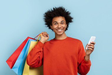 Günlük kıyafetli siyahi bir adam renkli alışveriş çantaları ve cep telefonuyla poz veriyor, tüketiciliği temsil ediyor, mavi arka planda.