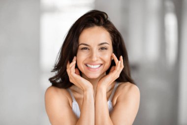 A joyful woman with a bright smile, holding her face with her hands, posed against a blurred background clipart