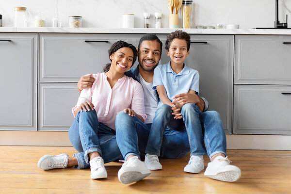 Black parents and child laughing together while seated on the kitchen floor, displaying a relaxed family atmosphere