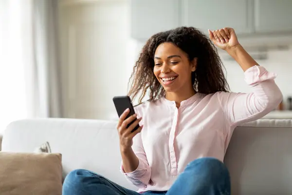 自宅にいるアフリカ系アメリカ人女性は おそらく彼女の携帯電話で良いニュースを受け取ることから喜びを表現し 興奮を体現しています — ストック写真