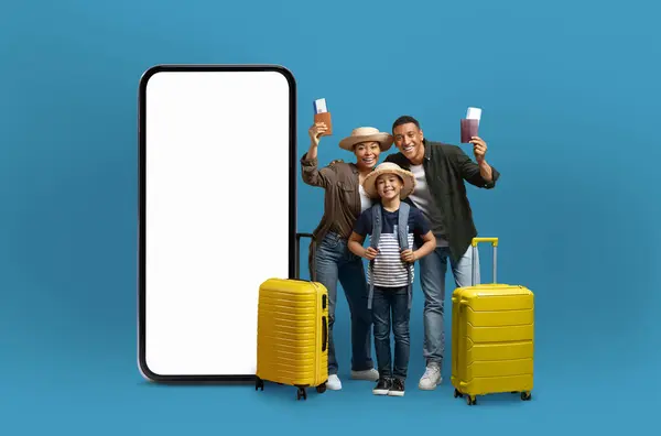 アフリカ系アメリカ人の家族は 巨大なスマートフォン画面の横にある旅行の必需品をポーズし 接続された旅行とデジタルアプリケーションを象徴しています — ストック写真