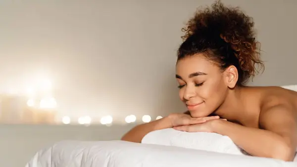 stock image This image captures a black woman lying down with a peaceful expression, symbolizing relaxation and self-care at a spa with people in the background