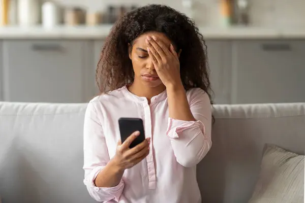 Stock image A photo capturing the tension of a young African American woman sitting on her couch, visibly overwhelmed as she looks at her smartphone in a neatly arranged living room