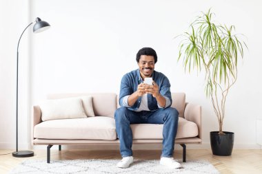 Neşeli Afrikalı Amerikalı adam evde akıllı telefonuyla mesaj atıyor, iletişim ve mutluluğu ifade ediyor.