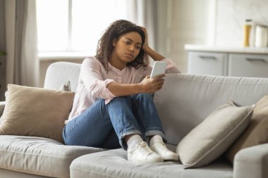 Afro-Amerikalı bir kadın telefonunu rahat bir oturma odasındaki koltukta kullanırken stresli ve endişeli görünüyor. Dijital iletişimin zorluklarını yansıtıyor.