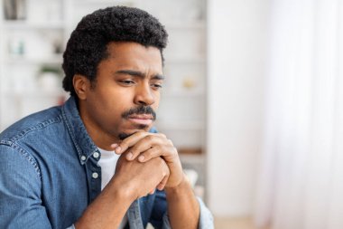 Düşünceli, Afro-Amerikan bir adam evde elleri sımsıkı bağlı, düşüncelere dalmış ya da endişeli bir şekilde bakıyor.