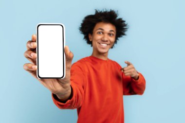 Turuncu kazaklı neşeli siyahi adam boş bir akıllı telefon ekranını tanıtıyor, içerik ekranı için uygun, mavi izole bir arka plan.