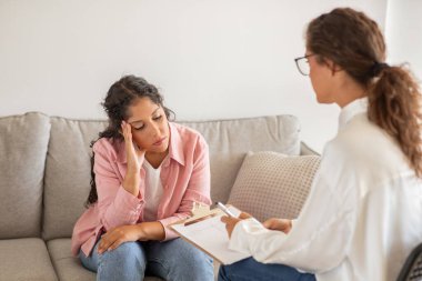 Endişeli bir bakışla genç bir kadın elinde bir dosya olan bir bayan danışmanı dikkatle dinliyor ve onunla konuşuyor..