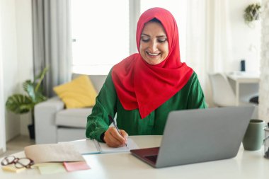 Müslüman kadın bir masada oturuyor ve dizüstü bilgisayarına konsantre oluyor. İşine ya da çalışmasına odaklanmış ve meşgul görünüyor. Oda aydınlık ve bayan duruşu profesyonel bir ortam öneriyor..