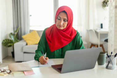 Tahta bir masada oturan tesettürlü bir kadın dizüstü bilgisayarına odaklanmış. Yanında bir fincan kahveyle çalışıyor ya da çalışıyor gibi görünüyor..