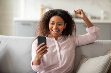 Huzurlu bir ev ortamını yakalayan Afro-Amerikan kadın, elinde akıllı telefonuyla koltuğa uzanmış rahat ve modern bir yaşam tarzını resmediyor.