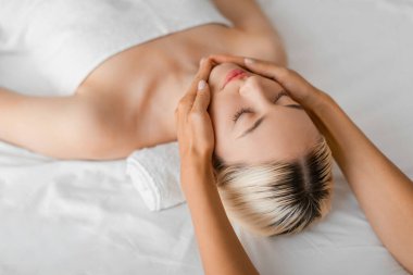 Bir kadın kapalı gözlerle uzanıyor, sakin bir kaplıca ortamında, yüksek açılı bir bakış açısıyla profesyonel bir terapistin rahatlatıcı yüz tedavisinin tadını çıkarıyor.