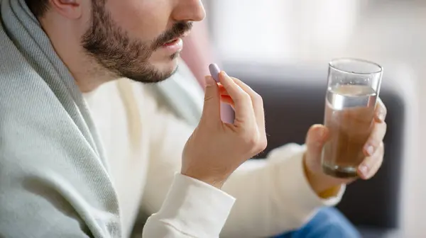 stock image Cropped of bearded man holding a pill between his fingers, ready to swallow it with the aid of a full glass of water, highlighting a moment of personal health care