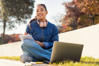 Bir kız öğrenci dışarıda oturuyor, dizüstü bilgisayarda çalışıyor, ödev yapıyor, kameraya gülümsüyor, boşluğu kopyalıyor.