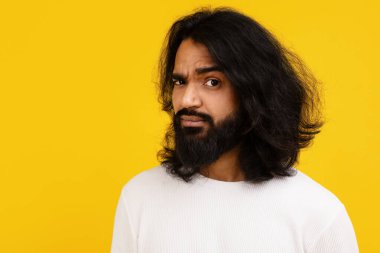 A perplexed young Indian man with a furrowed brow stands against a vivid yellow backdrop, exhibiting a puzzled expression, closeup clipart