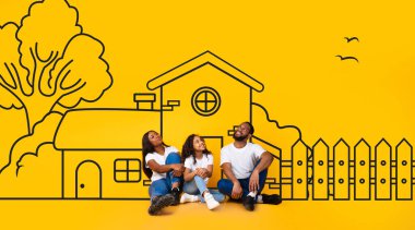 Üç çocuklu mutlu siyah aile, anne ve kız yerde oturuyor ve sarı duvar arkasından hayallerinin resimli evini izliyorlar.