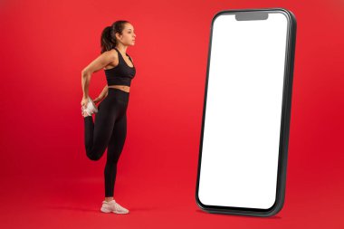 Siyah spor elbiseli bir kadın, bir bacağını kaldırıyor, arkasında, büyük bir büyük, dikey bir akıllı telefon yanında boş bir ekran, canlı kırmızı bir arka plan karşı